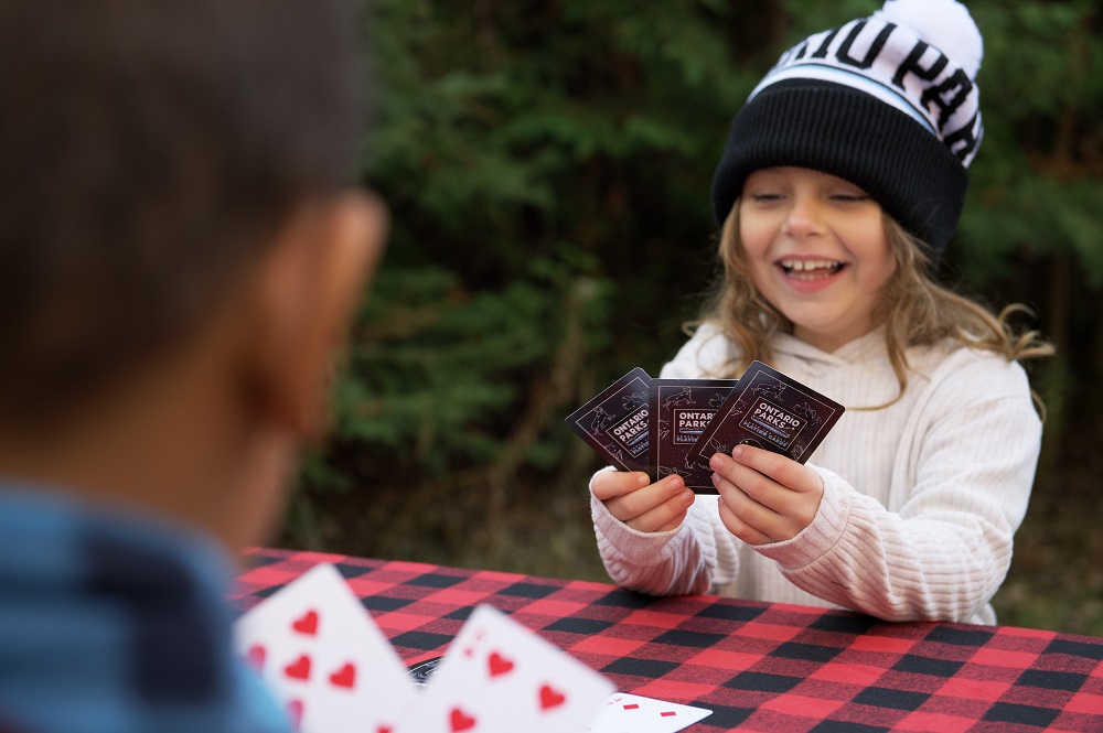 child sitting at table with buffalo plaid tablecloth, wearing toque and holding cards