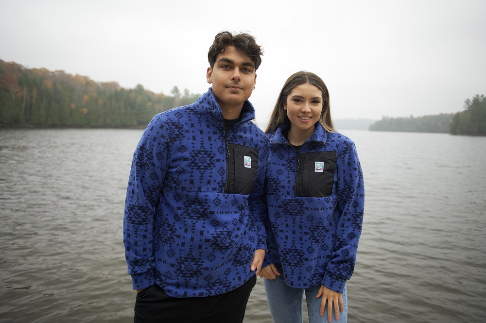 two models wearing blue quarter zip sweaters, standing in front of lake