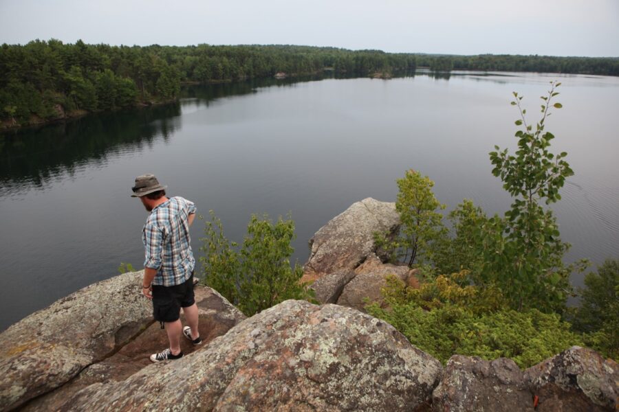 A person standing on a rockface lookout over a large lake with a treed shore in the distance
