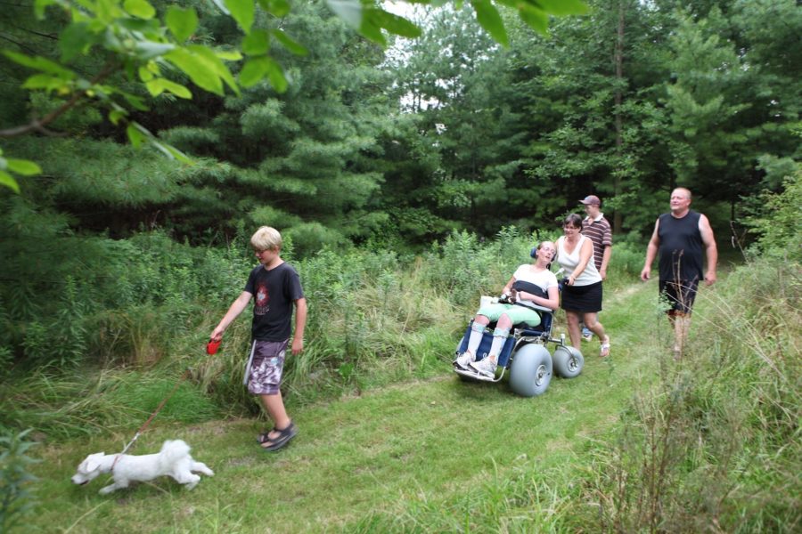 5 people, including one person in a wheel chair built for trails, and a dog, on a hike on a grassy trail through the woods. 