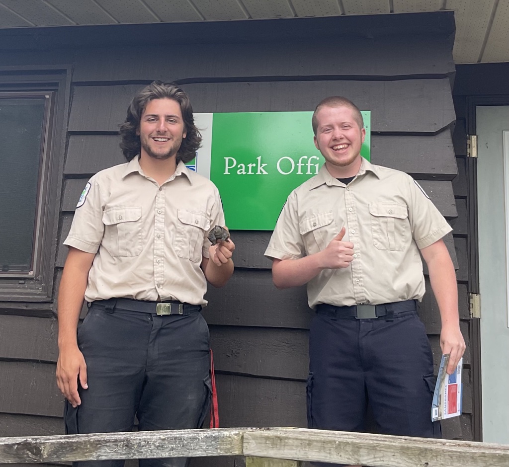 Deux employés de Parcs Ontario portant leurs uniformes beige et noir, debout devant un bâtiment avec un panneau indiquant « Bureau du parc ».