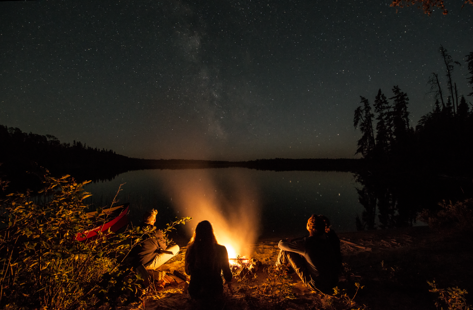 Group sits around campfire at night time by lake.