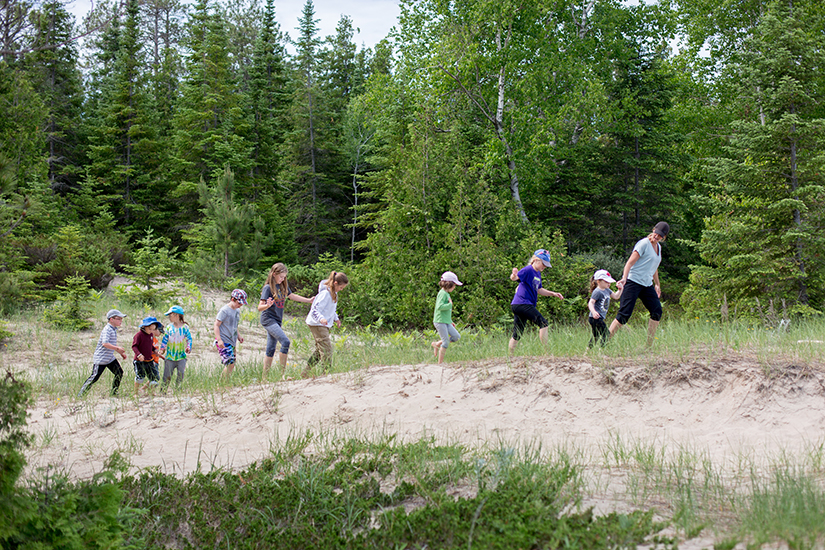 Children follow teacher across grassy beach area