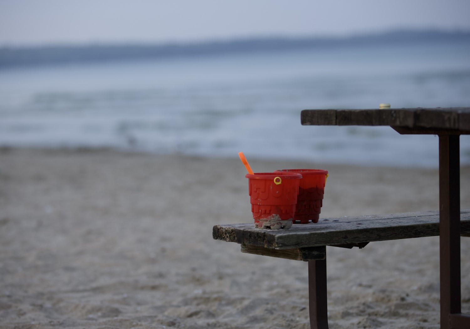 Two red sand buckets, children's beach toys, sitting on a wooden picnic table on a beach