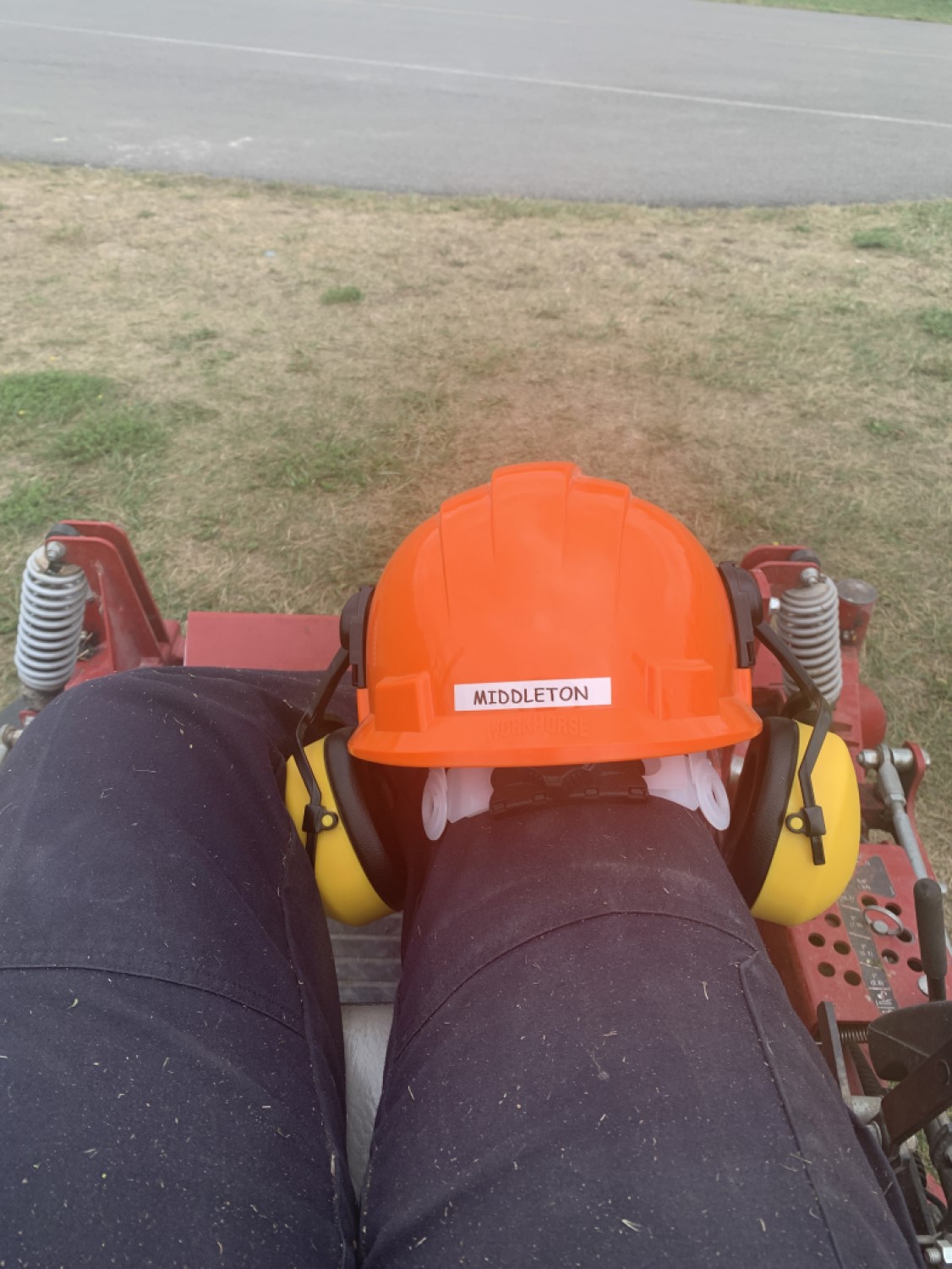 A person's legs with their orange hardhat and ear protection resting on their right knee. They are sitting in some type of small machine, like a riding lawnmower.