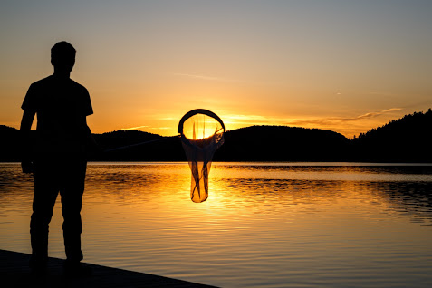 Silhouette of a person holding a bug net standing on a dock as the sun sets in the distance.
