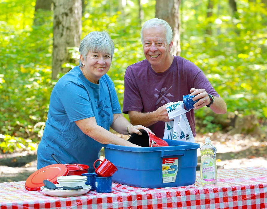 Couple with grey hair washing colourful enamel dishes in a blue container on a picnic table with a red and white checked table cloth