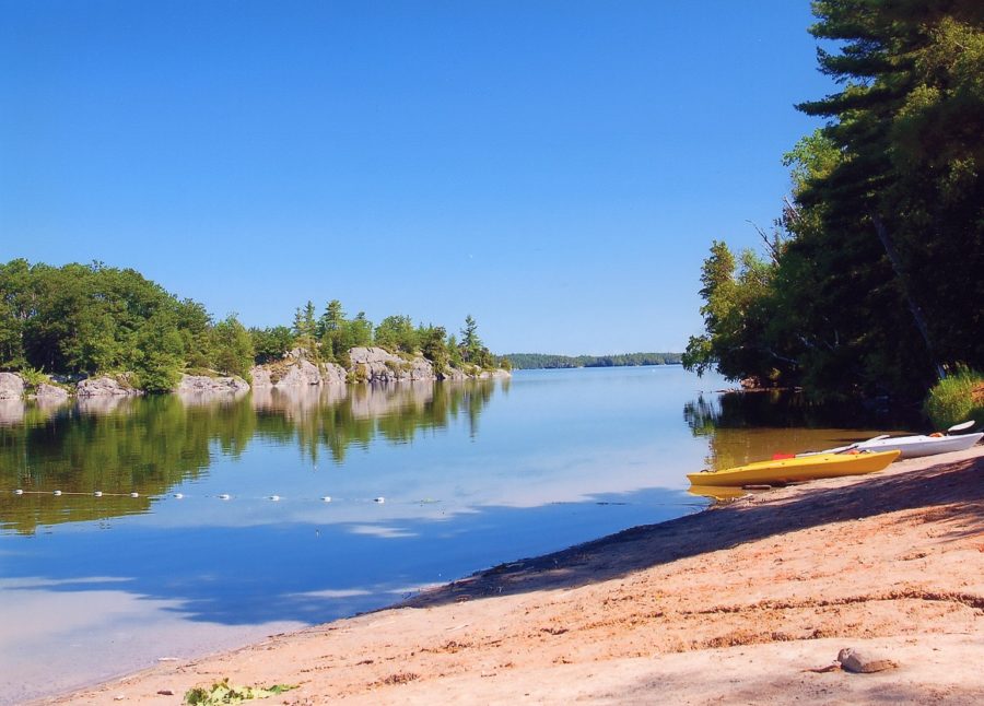 A sunny day with a beach in the foreground, a lake with a swimming area just beyond and a rocky shoreline in the background. Two kayaks are on the beach - 1 yellow, and 1 white. 
