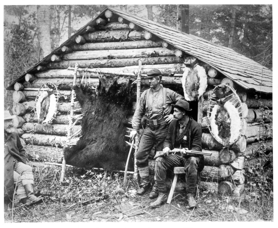 Two men outside a primitive cabin circa 1897. Men are surrounded by various hides being stretched. 