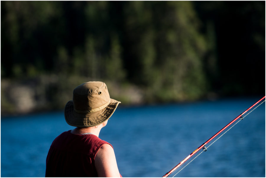 Une personne vue de dos coiffée d’un chapeau de randonnée qui pêche au soleil. L’arrière-plan consiste en un lac bleu et une ligne de rivage verte.
