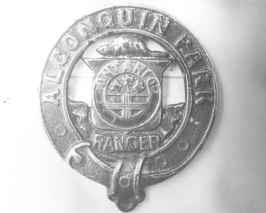 Black and white image of a circular brass pin, including the Ontario crest, that says Algonquin Park Ranger. 