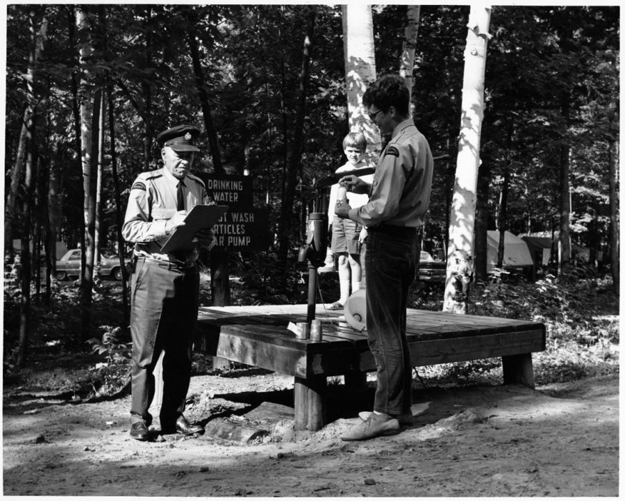 Black and white image. Two male park staff sampling water and recording, stand in front of a platform. In the background stands a little kid watching on.