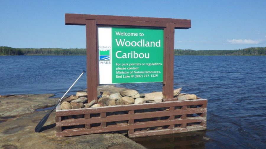 A Ontario Parks entrance sign on a lake, that says "Welcome to Woodland Caribou, for park permits or regulations please contact Ministry of Natural Resources, Red lake at 807 727 1329