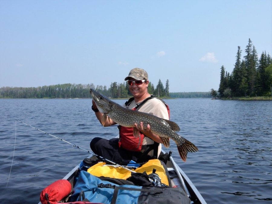 Fisherman in the front of a packed canoe, holding up a 2.5 ft long Northern Pike