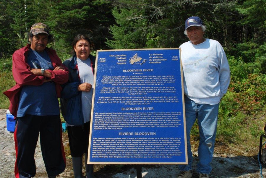 A group of two men and a woman standing around a plaque that is titled Bloodvein River. These people are Anishinaabek and were born and have lived on the Bloodvein River. 