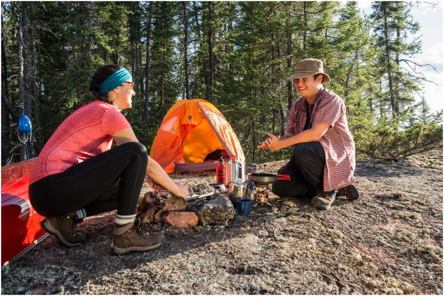 Two people cooking at a campsite, with coniferous forest and an orange tent in the background