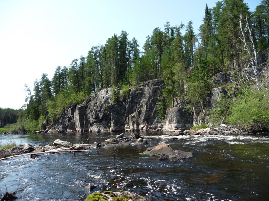 Vue d’un côté de la rivière – d’une dizaine de mètres de large – d’où l’on aperçoit d’imposantes formations rocheuses de trois mètres du haut desquelles poussent des conifères.