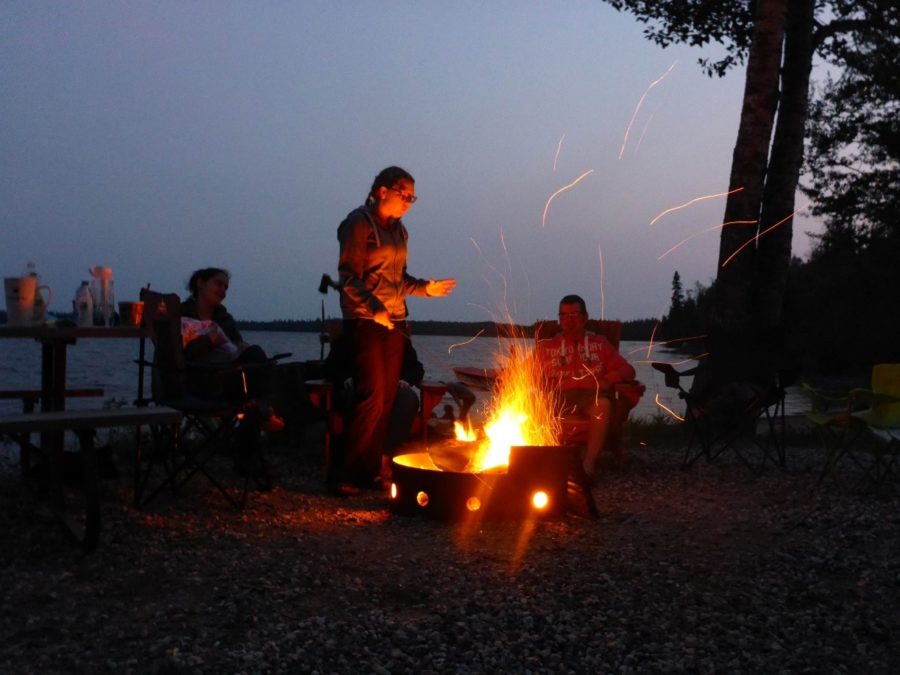 Three people at dusk around a campfire, one person is standing. 