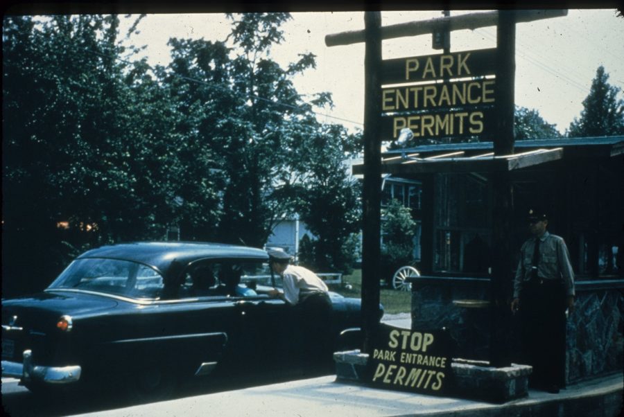 Park Entrance from the 50's or 60's with two park wardens in the shot, one of whom is speaking into the passenger side of a car. Visible signs say "Park Entrance Permits" and "STOP Park Entrance PERMITS"