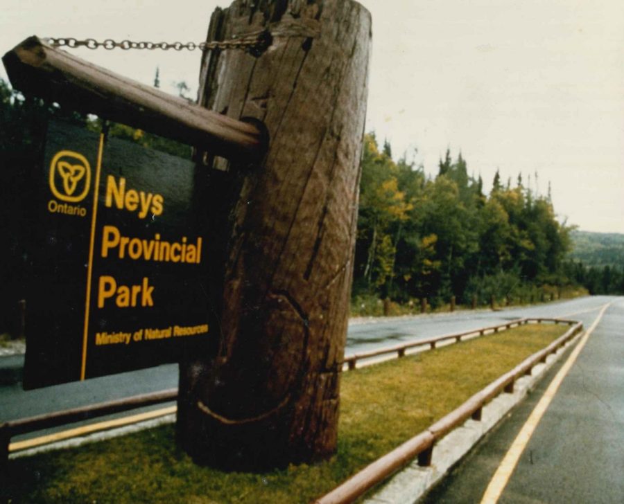 Photo granuleuse d’un panneau de signalisation brun foncé portant le logo trifolié jaune de l’Ontario et l’inscription jaune « Neys Provincial Park » -- accroché à un grand rondin massif.