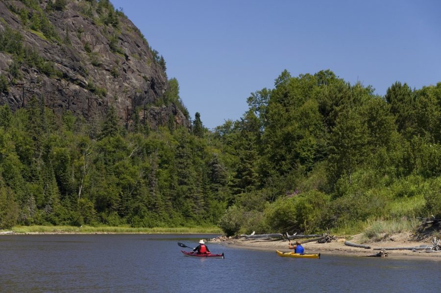 Two kayakers on a sunny day with rock feature in front of them, paddling alongside a quiet shoreline with brush and forest