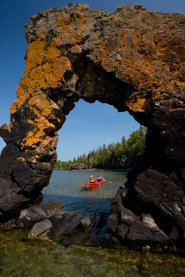 Formation rocheuse percée d’un trou, avec du lichen jaune brillant, émergeant de l’eau. Par le trou, vous pouvez apercevoir deux personnes dans un kayak.