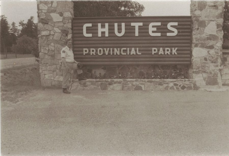 Photo en noir et blanc d’un panneau de signalisation foncé indiquant Chutes Provincial Park, sans logo, flanqué de piliers de pierre et comprenant un garde de parc en arrière-plan.