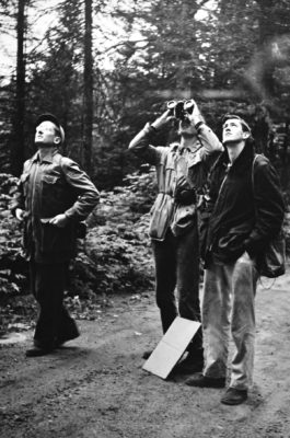 Photographie en noir et blanc de trois hommes scrutant les hauteurs (dont un avec des jumelles).