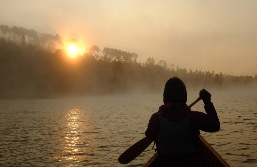 View from the back of a canoe on the water of sillouette of person paddling in the front and the sun is peeking over trees ahead