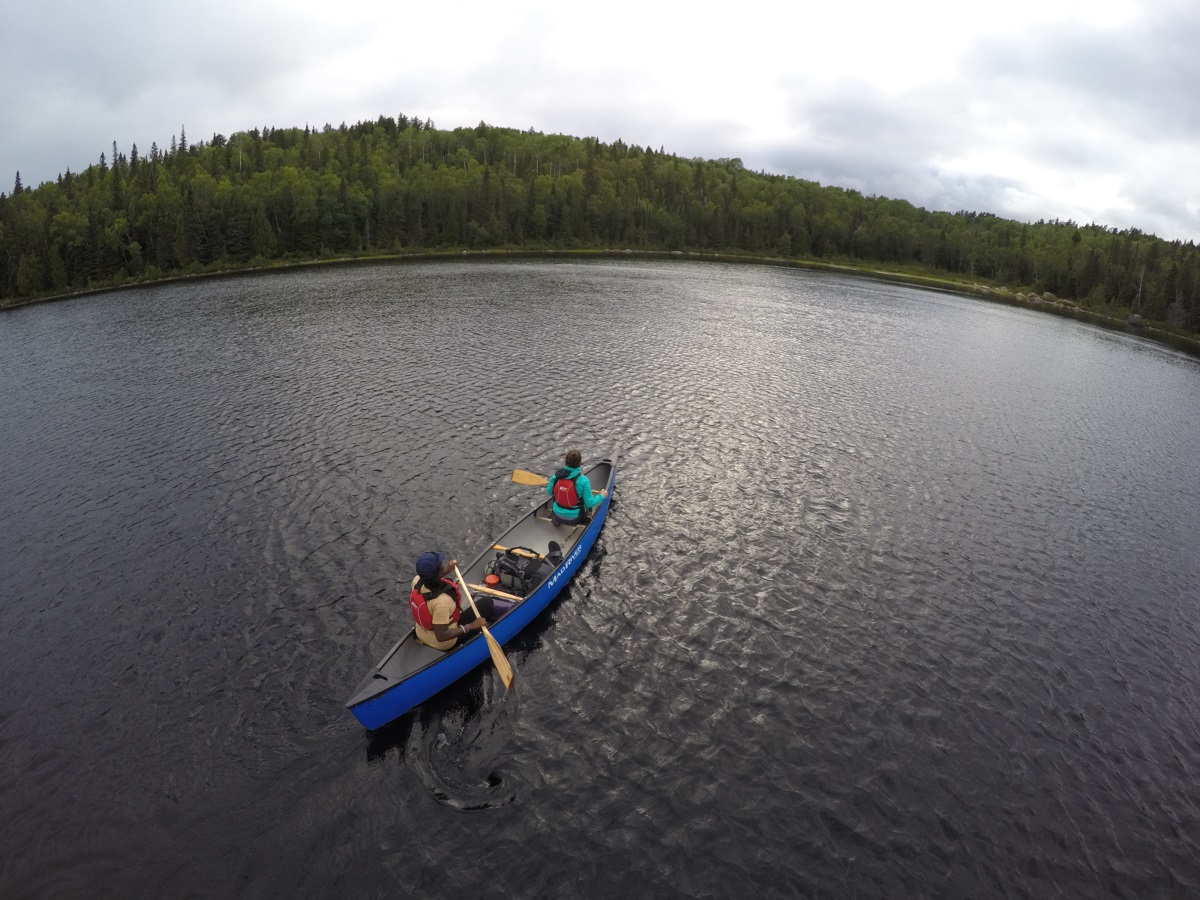 Two people paddling blue canoe over lake towards forested shoreline on a grey day