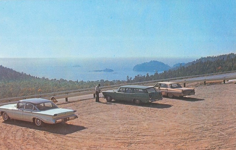 Three cars from the 60s parked at a look out overlooking a large with a series of far off islands, and forest.