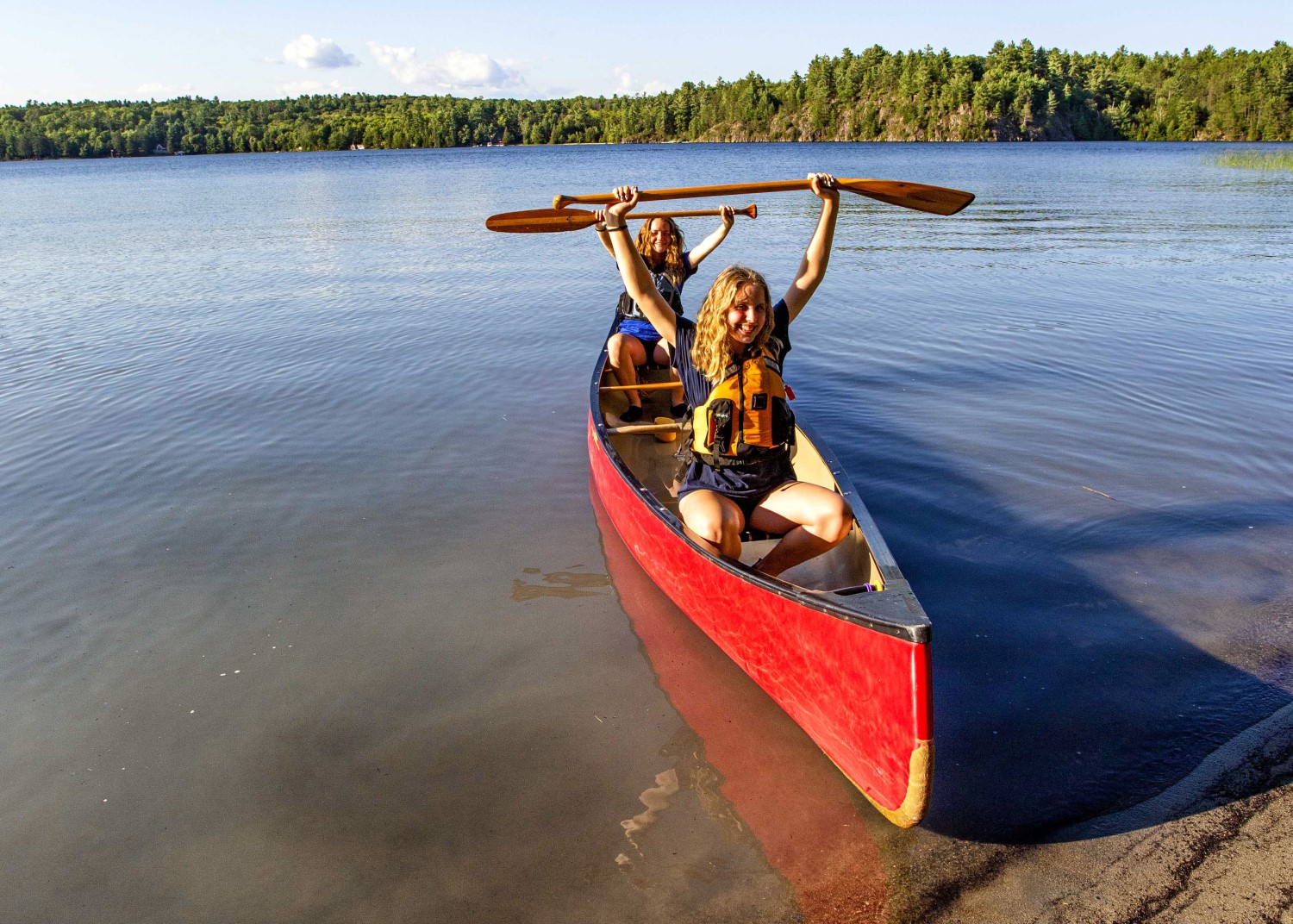 Two people sitting in a red canoe in the water, holding their paddles over their heads and smiling at the camera
