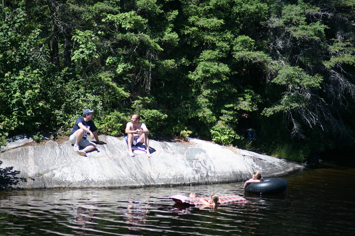 Two people sit on a rock shore facing the water while two other people relax in the river