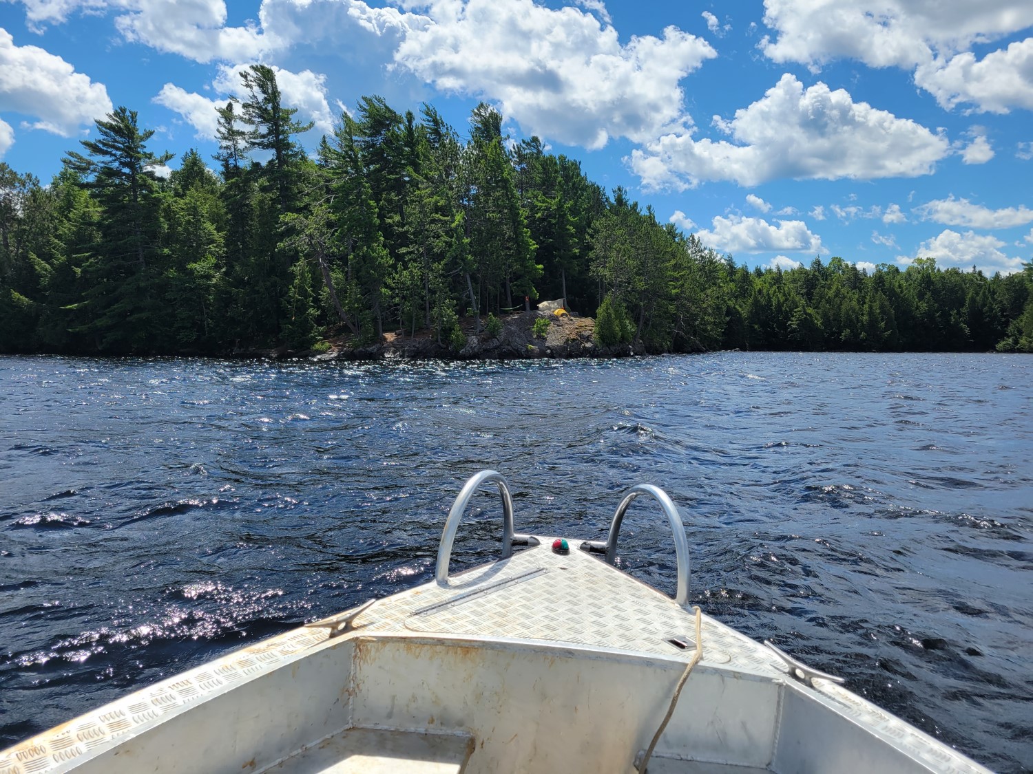 View from the front of a small metal boat facing toward a small, tree-covered island. In the clearing on the island, there is a small yellow tent.