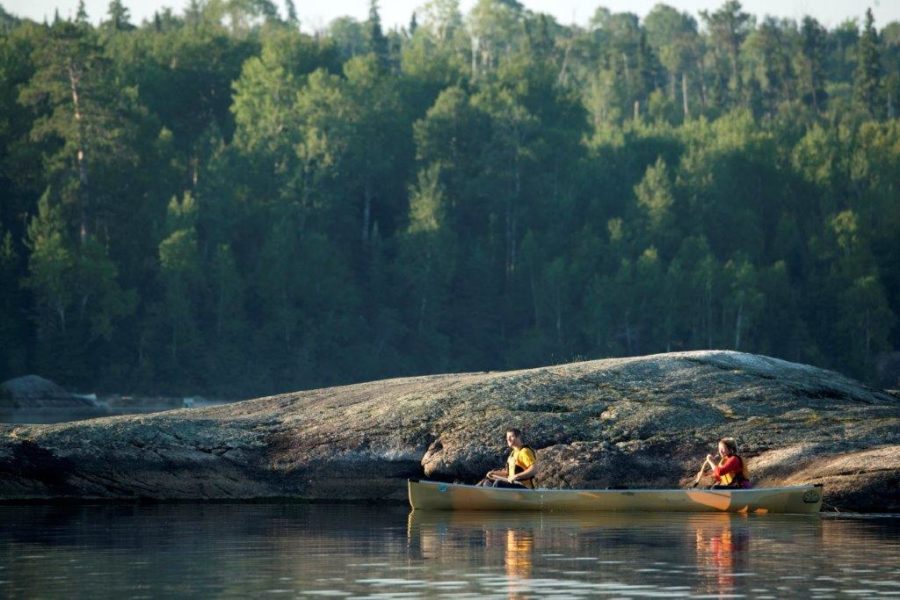 Deux pagayeurs à bord d’un canot, près d’un rivage rocheux avec une forêt en arrière-plan 