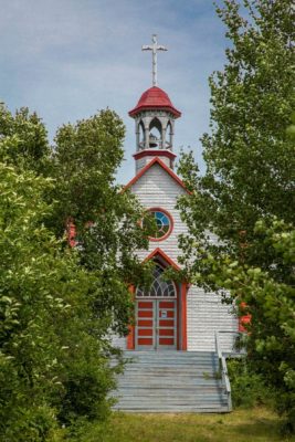 Old white church with bell tower, all with red trim, behind some crowding deciduous trees