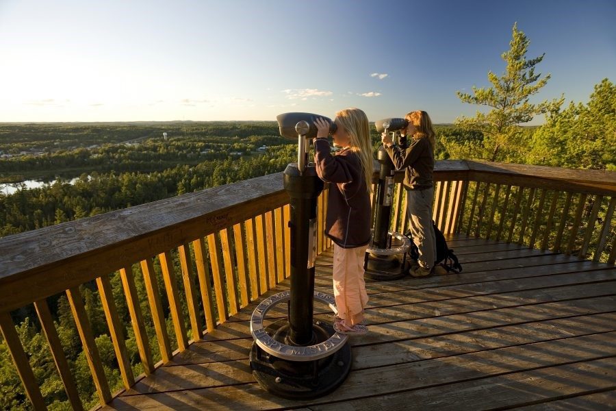 Two people look through stationary binoculars at a view of lakes and forests when the sun in low in the sky