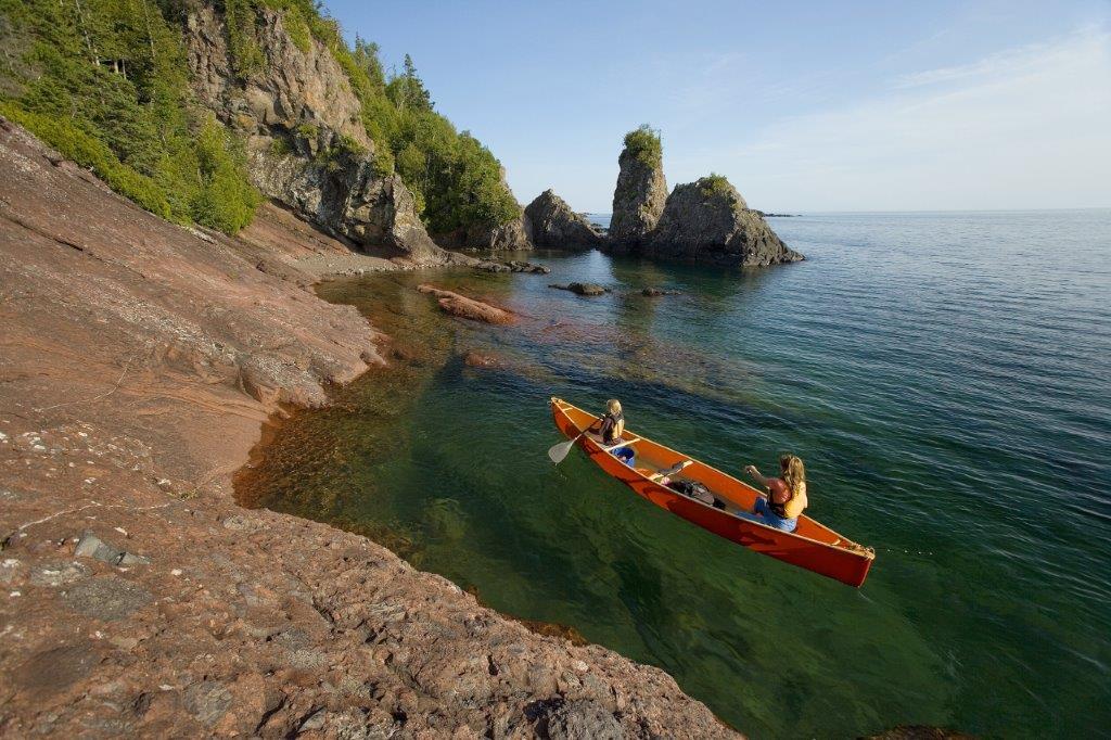 Red kayak approaching a shore with reddish rocks on a sunny day
