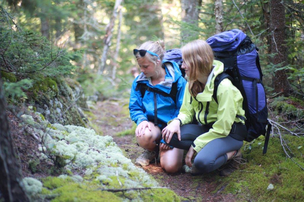 Two girls with big hiking backpacks crouching down to look at moss in the forest
