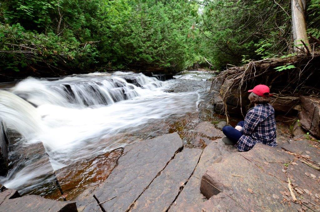 Person in a red cap sitting next to a rushing river that is lined with ceder trees