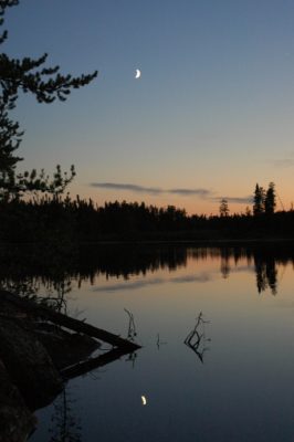 Moon at sunset on a reflective lake with silhouetted forest