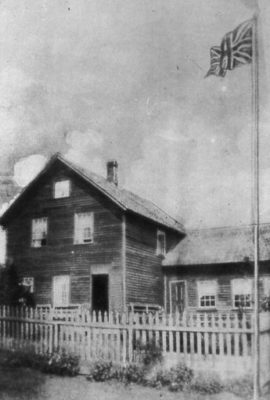 Black and white image of dark two story structure with flag flying out front