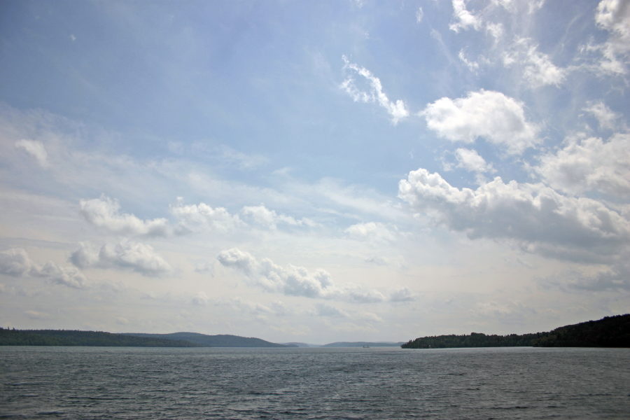 Lac sous un ciel bleu avec des nuages blancs cotonneux et forêt loin à l'horizon.
