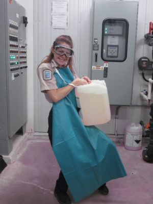 Girl in an industrial room, wearing a protective smock and holding up a plastic container filled with unknown liquid