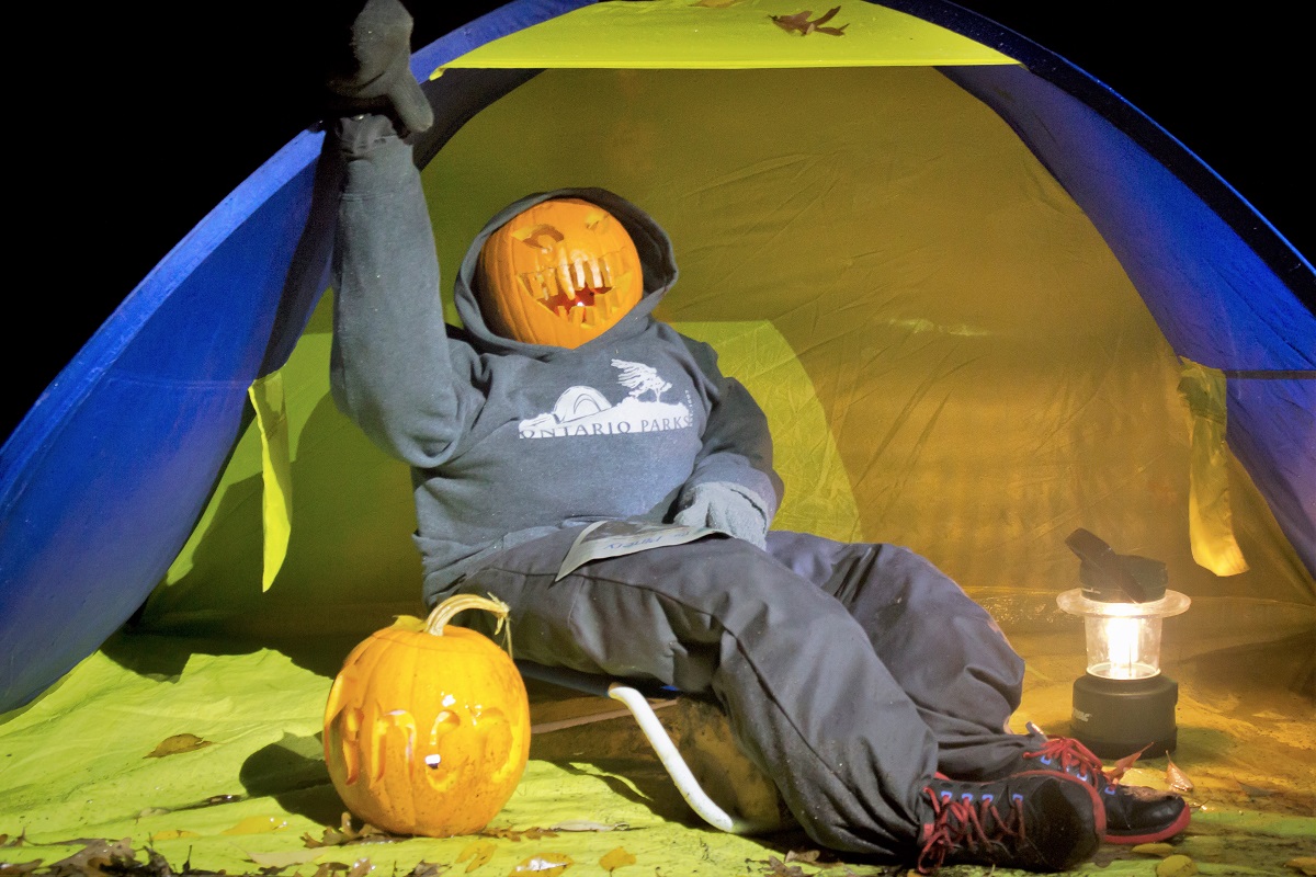 Un homme avec une tête en citrouille-lanterne, assis sur une chaise de camping devant une tente