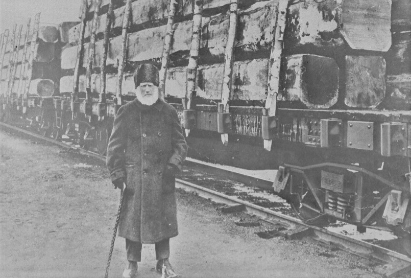 Black and white photo of older guy standing in front of train cars with railway ties