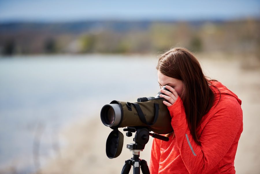 Une ornithologue amateur sur la plage. Elle porte une veste à capuche rouge et regarde à travers un télescope. 