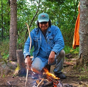 Homme dans les bois, tenant un bâton, accroupi devant un feu.