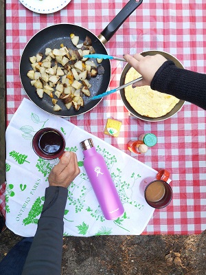picnic table with plaid cloth. frying pan with potatoes and water bottle and mug.