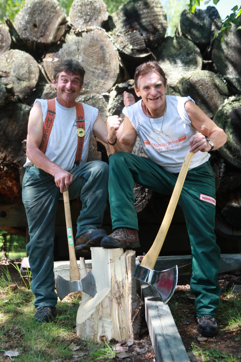 Two people wearing red suspenders and green pants, each with one leg up on a piece of chopped wood. The are both leaning against the handles of axes.